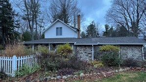A house with a stone foundation and a chimney, surrounded by a white picket fence and various plants. - Torch Lake Farmhouse (Alden)