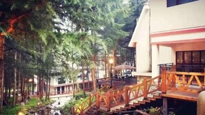 A wooden bridge over a stream, surrounded by trees and a building with a balcony.