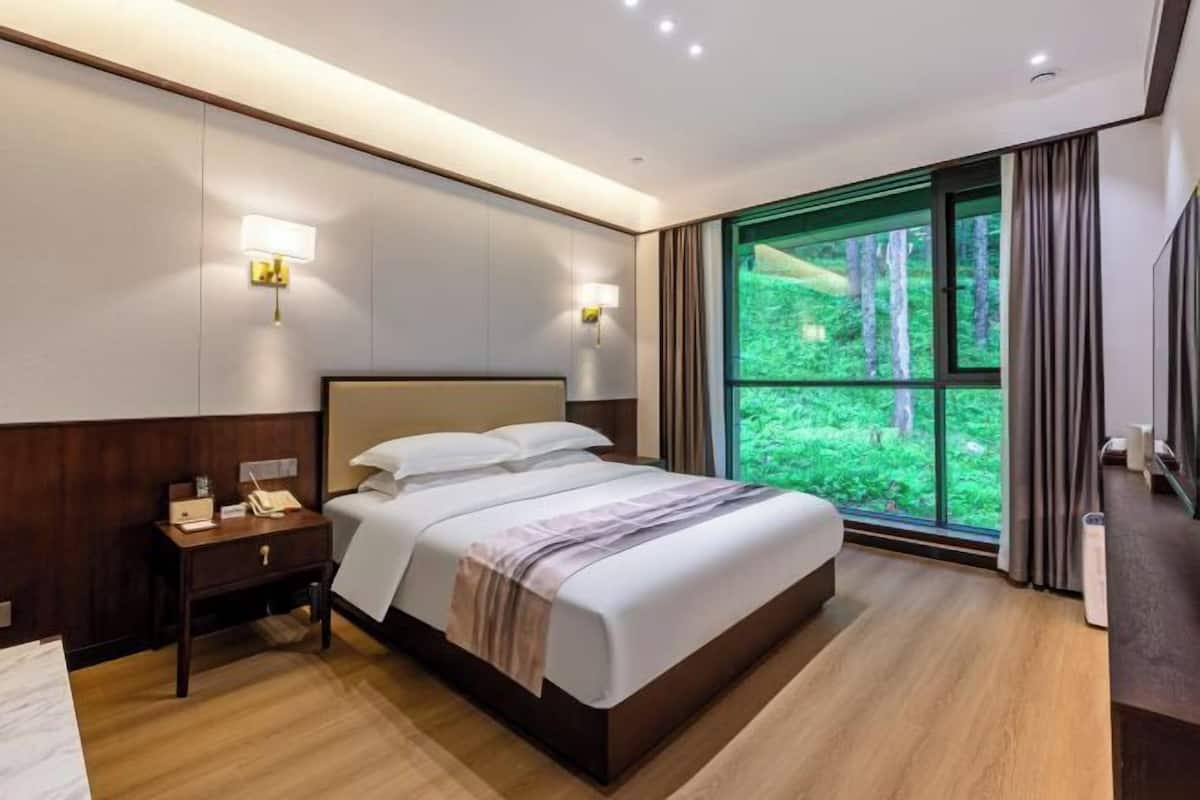 Una habitación de hotel moderna con una cama grande, mesitas de noche y vistas a un bosque a través de un amplio ventanal.