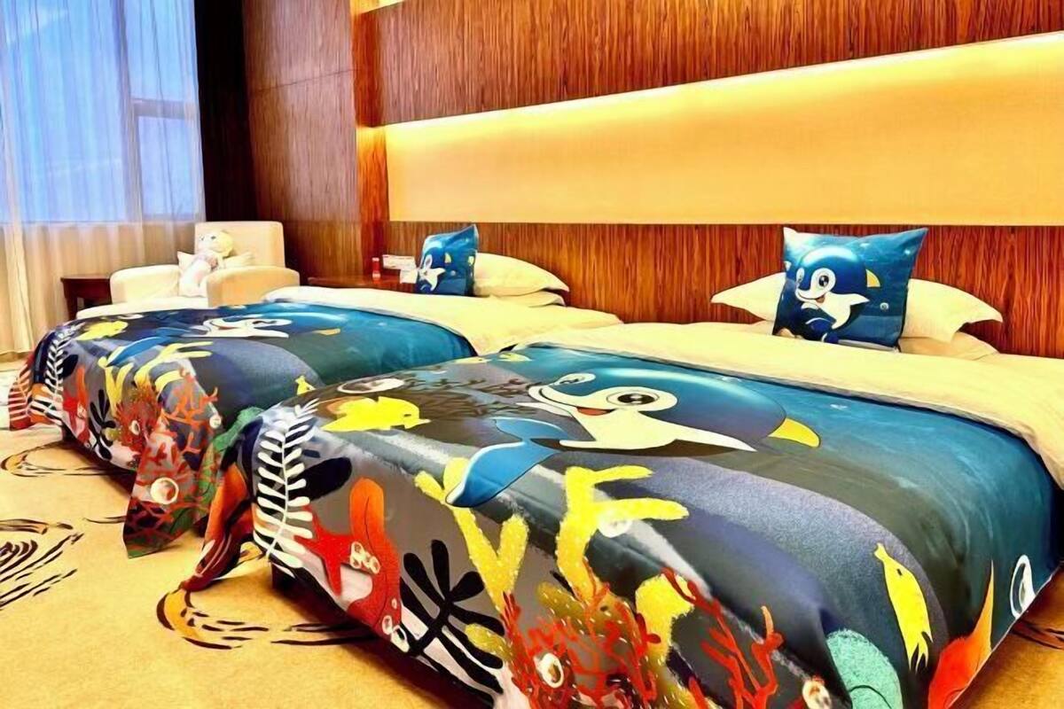 Habitación de hotel con dos camas, cada una con ropa de cama colorida adornada con personajes de caricaturas. Cuenta con un cabecero de madera y una pequeña zona de estar con una silla y un oso de peluche.