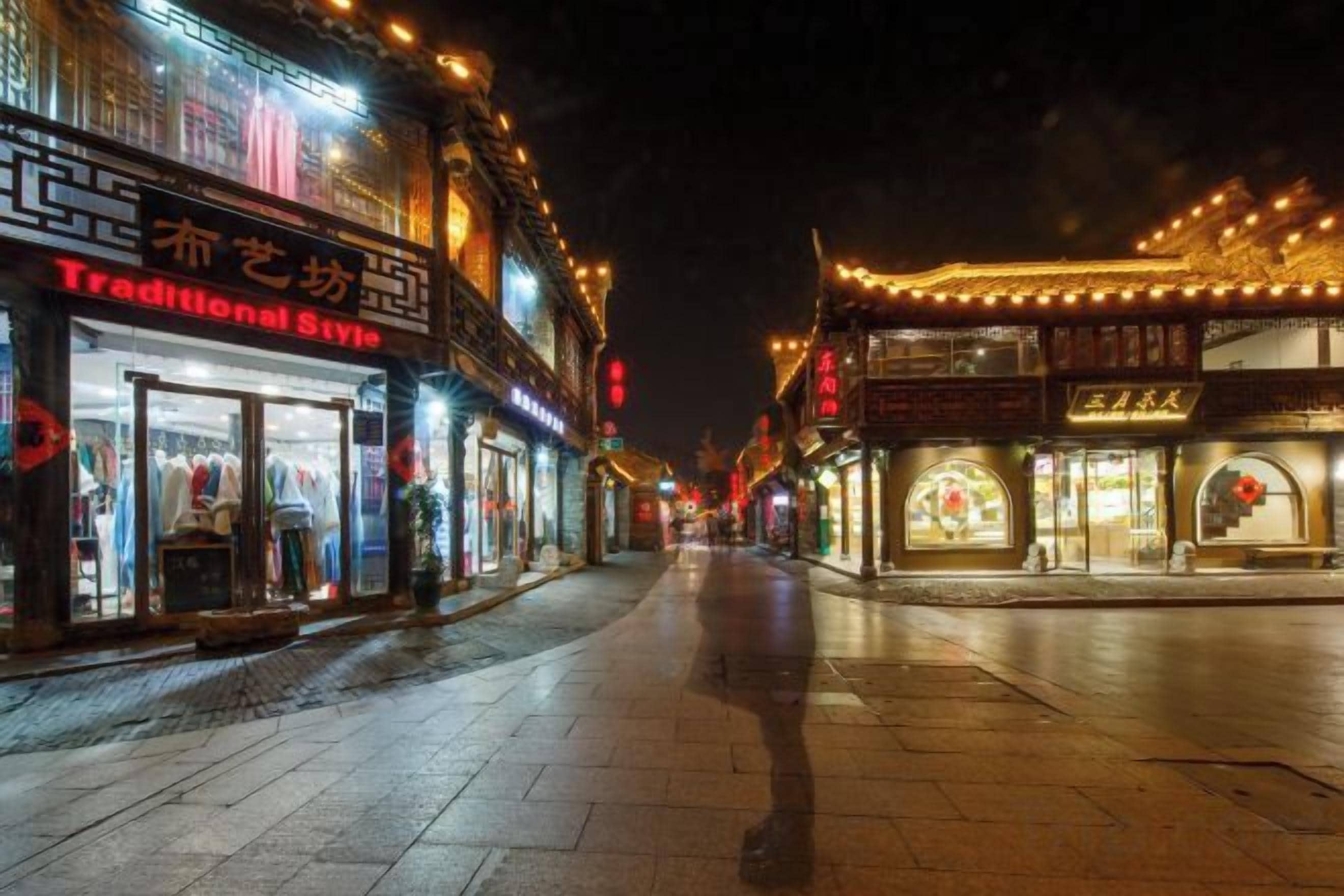 A nighttime street scene with illuminated storefronts and traditional architecture.