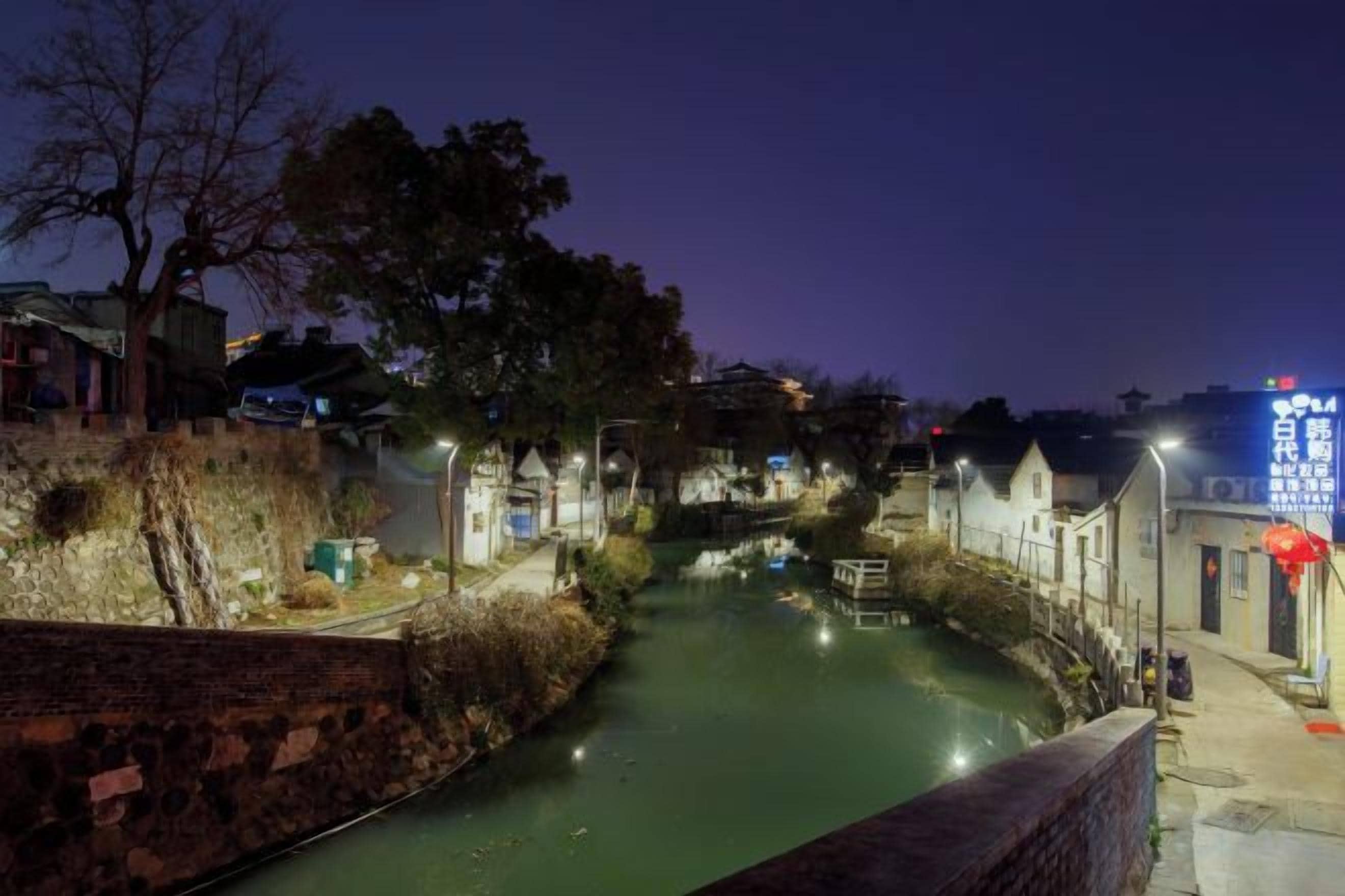 Une vue nocturne d’un étroit canal bordé de bâtiments dont les fenêtres sont illuminées, d’un mur de briques et d’un parapluie rouge.