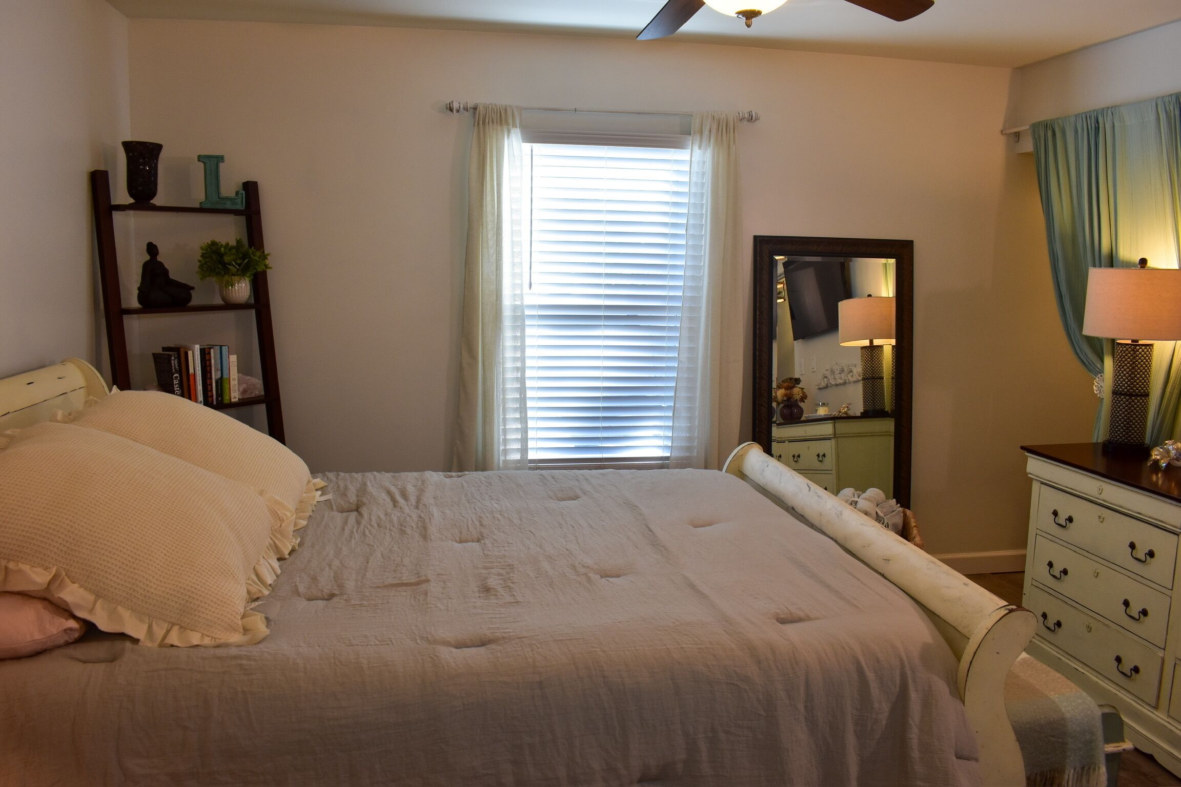 A bedroom with a bed, a dresser, a mirror, and a shelf with books and decorative items.