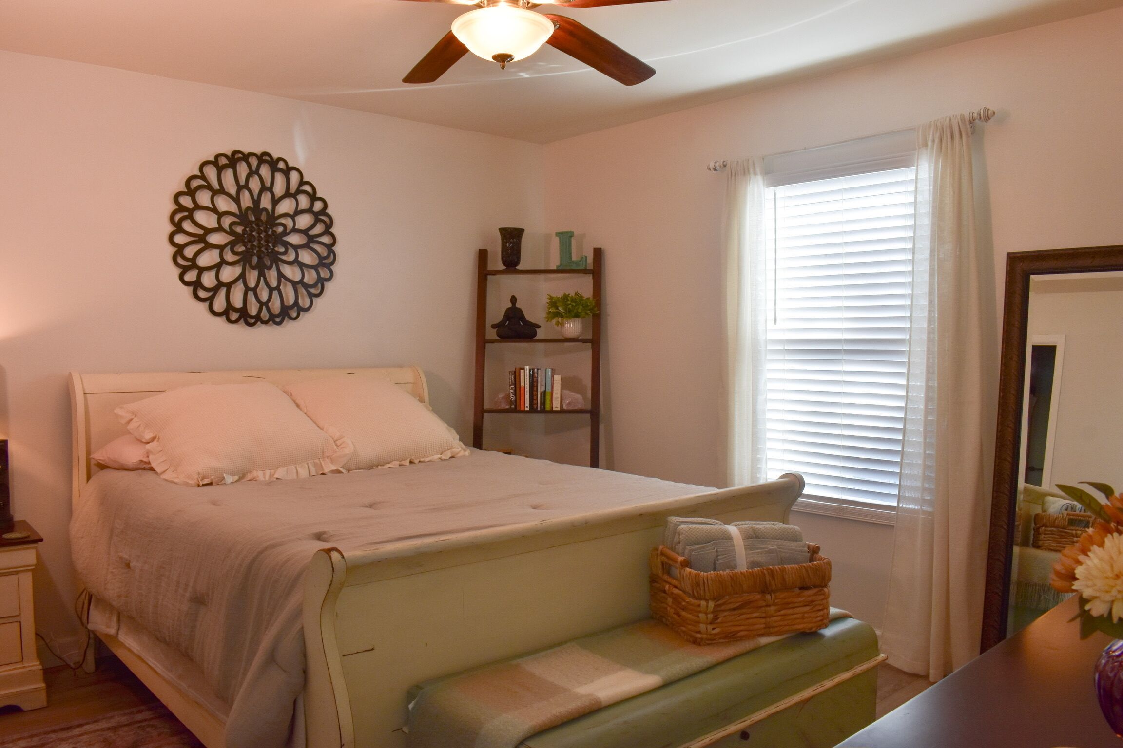 Une chambre à coucher avec un lit, une table de chevet, une étagère avec des livres et une plante, une fenêtre avec des rideaux et un ventilateur de plafond.