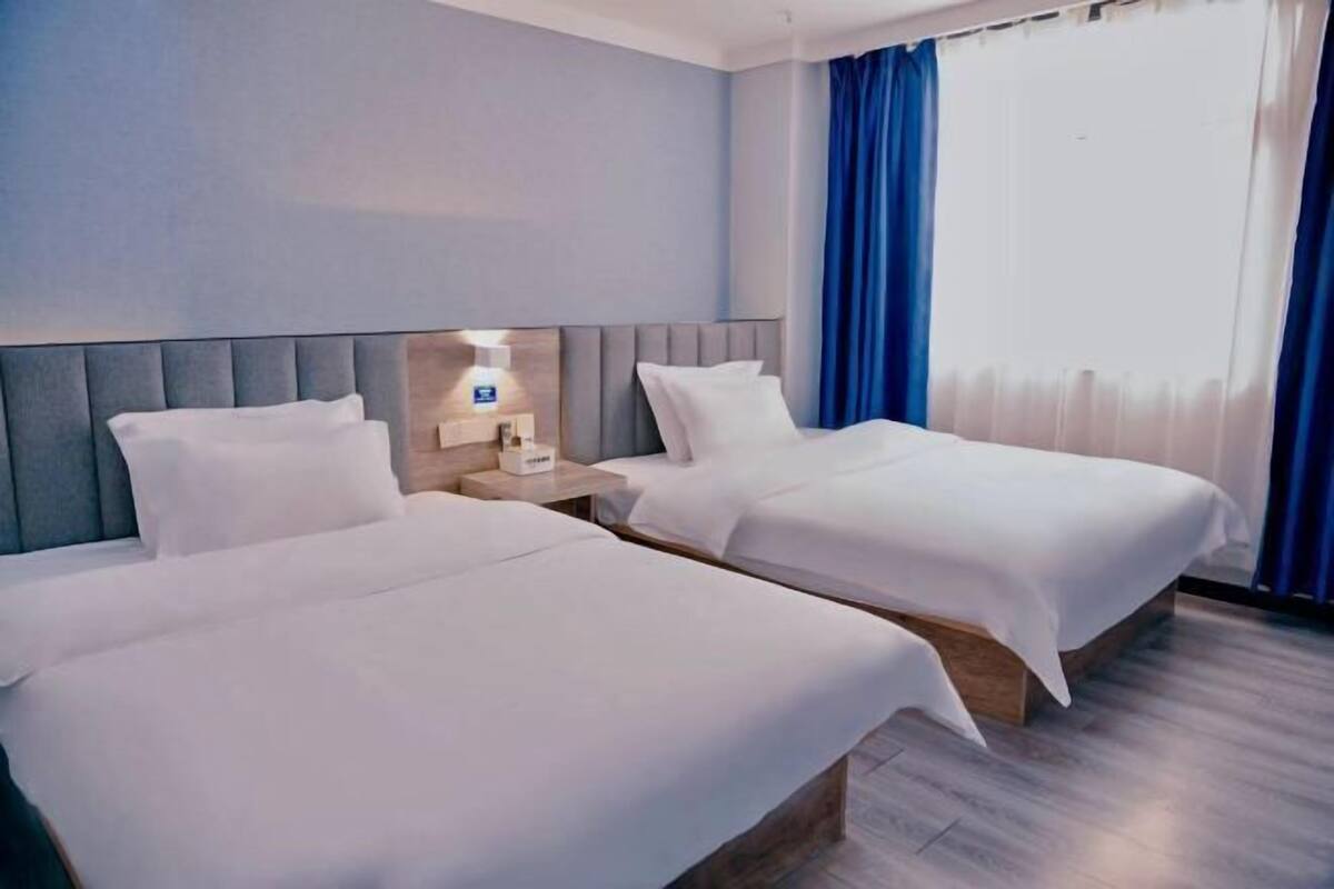 A hotel room with two single beds, a nightstand with a lamp, and a large window with blue curtains.