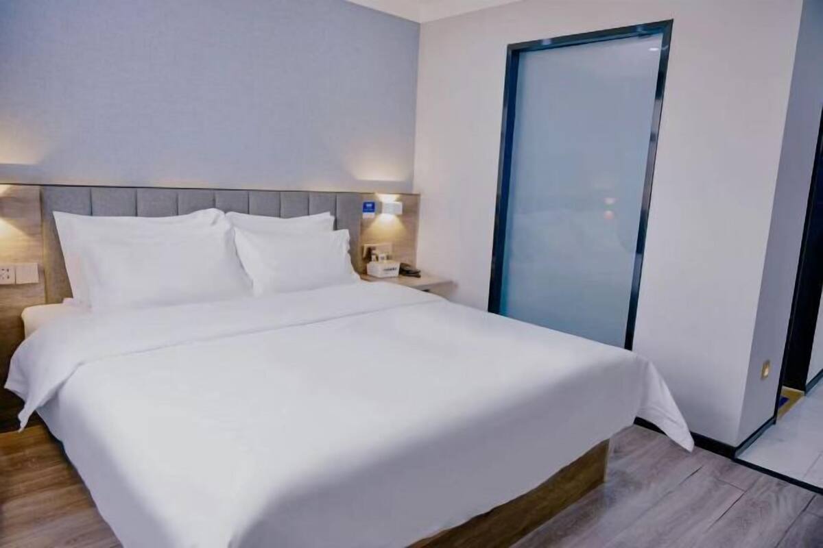 A hotel room with a large bed, a nightstand, a wall-mounted lamp, and a door.