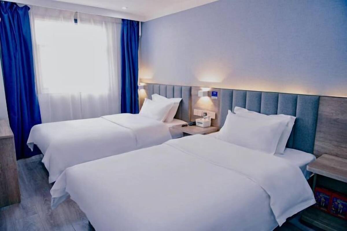 A hotel room with two beds, a wooden nightstand, a lamp, and a window with blue curtains.