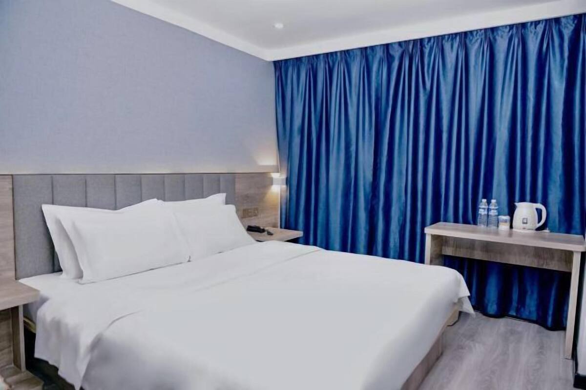A hotel room with a bed, a nightstand, a table with a kettle, and blue curtains.