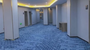 A hotel corridor with a patterned floor, recessed lighting, and a central hallway leading to a reception area. - 7 Days Premium (Zibo Zichuan Hongtai) (Zibo)