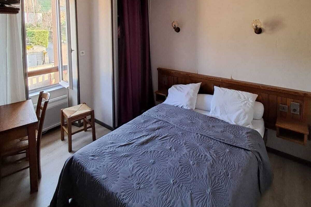 A hotel room with a bed, a desk, and a chair. There is a window with curtains, and a door is visible.