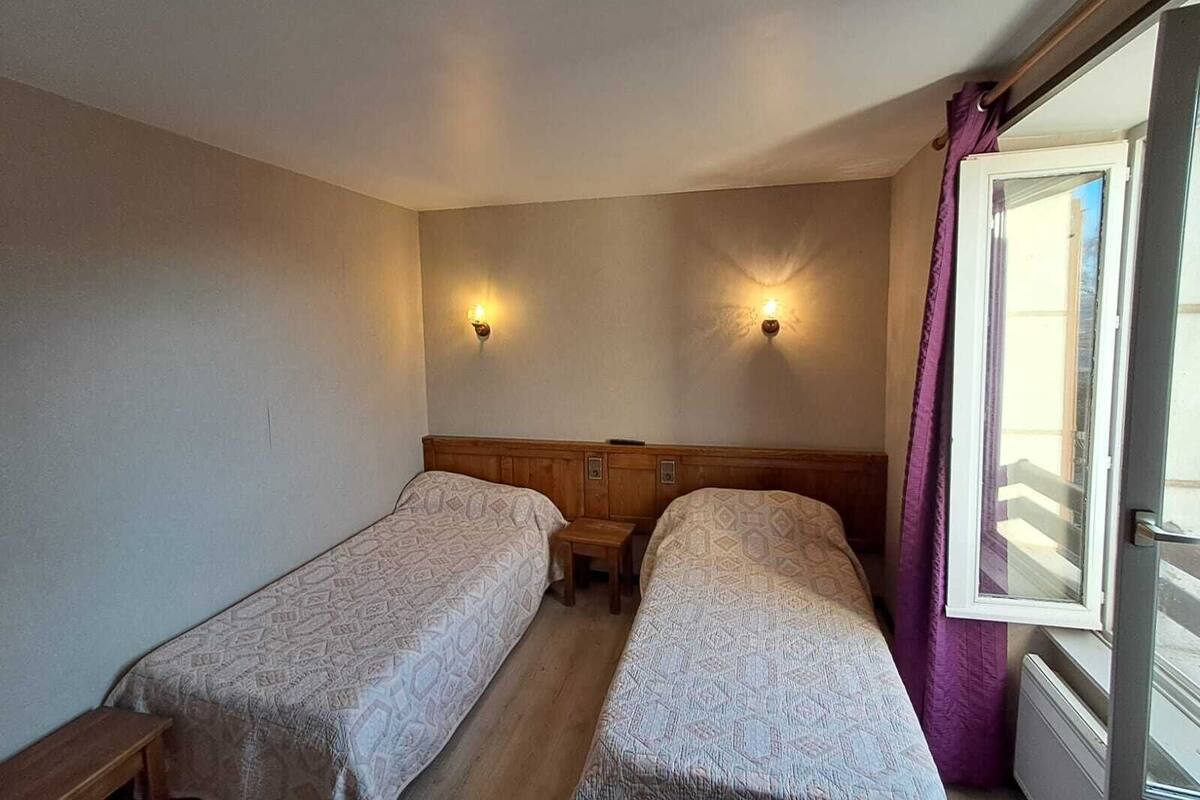 A small, simple room with two single beds, a small table, and a window with curtains.