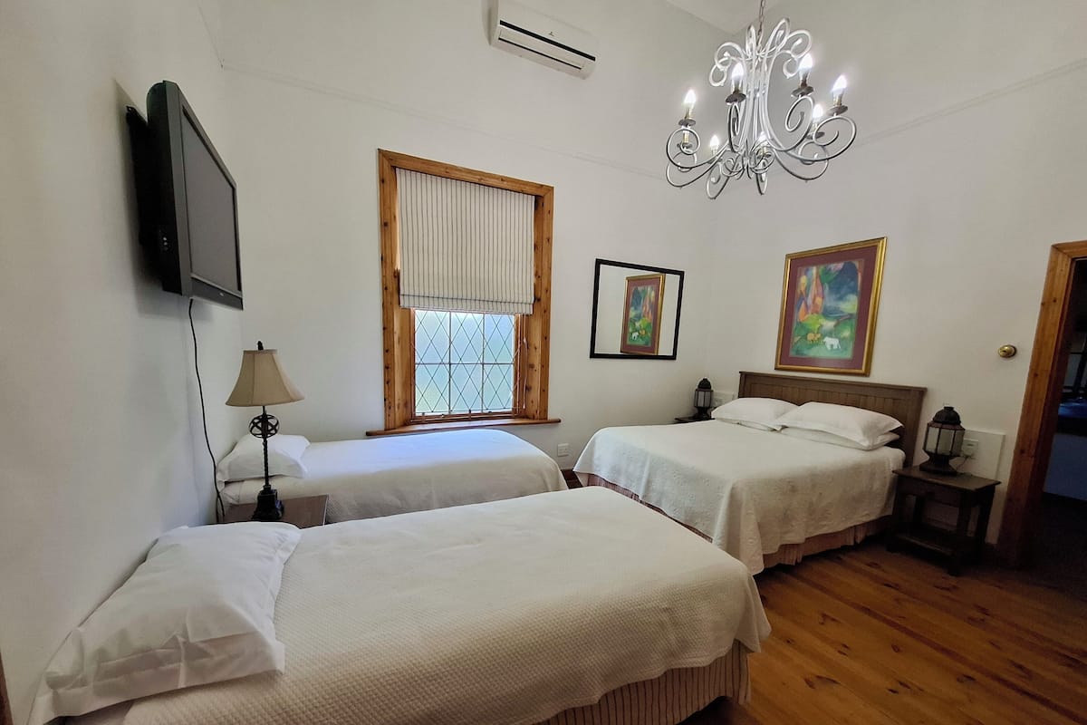 A hotel room with two beds, a TV, a window with blinds, a chandelier, and artwork on the walls.