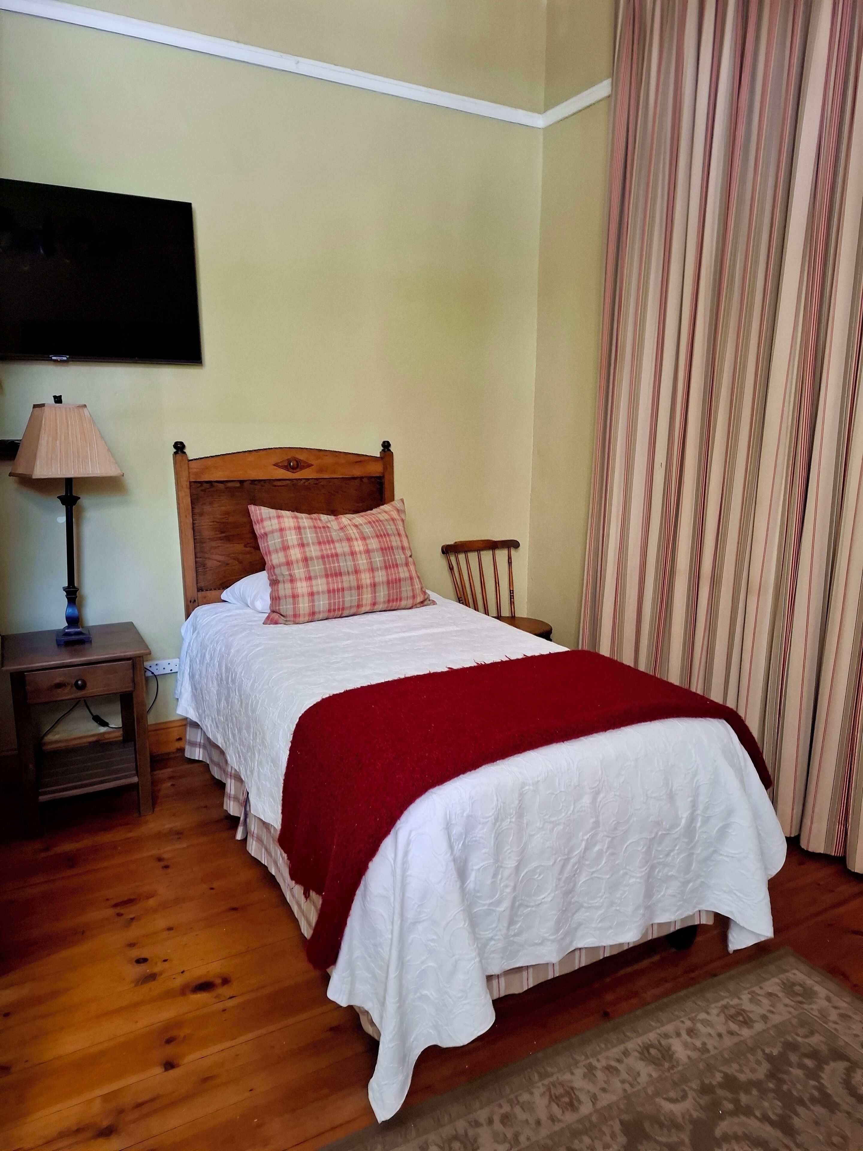 A neatly made bed with a wooden headboard, a red throw blanket, and a plaid pillow. There is a nightstand with a lamp beside the bed, and a wooden chair next to it.