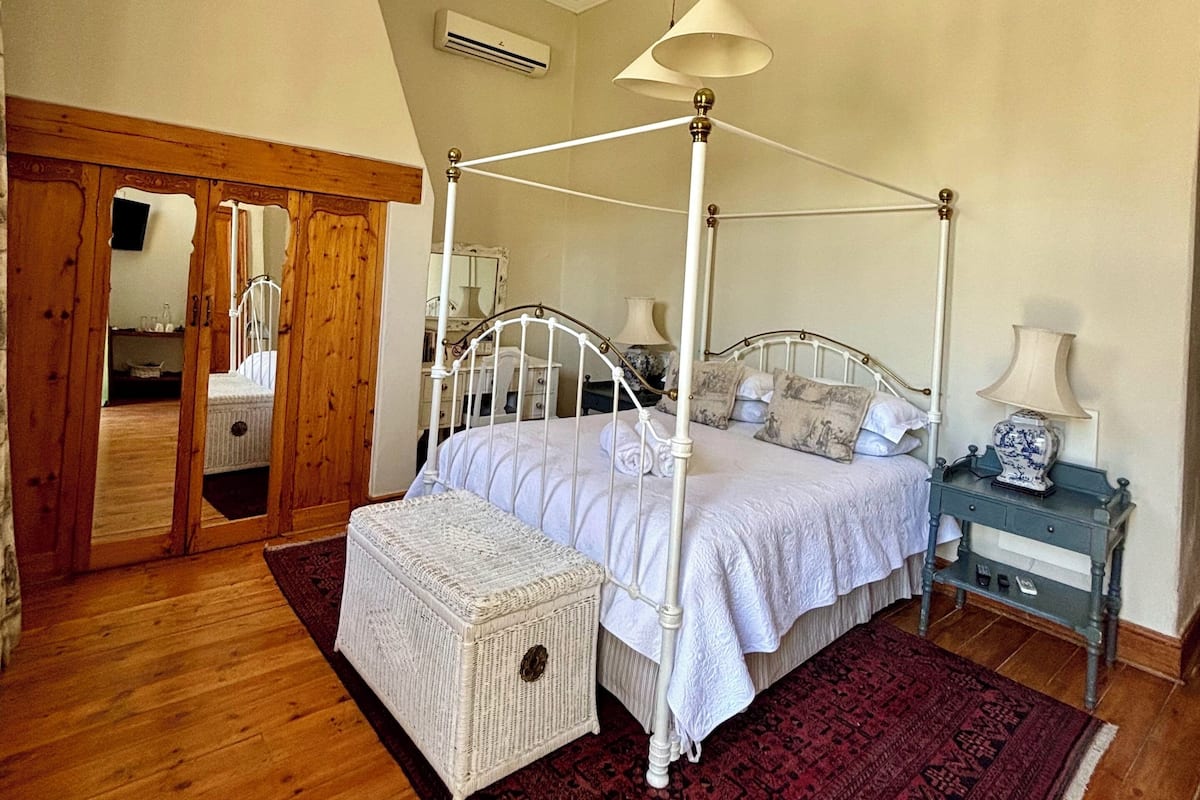 A four-poster bed with a hanging lamp, a bedside table with a lamp, a red patterned rug, and a wicker chest of drawers.