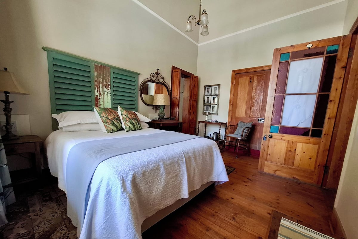 A bedroom with a large bed, wooden floor, a wooden door with glass panels, a small table with a lamp, and a mirror on the wall.
