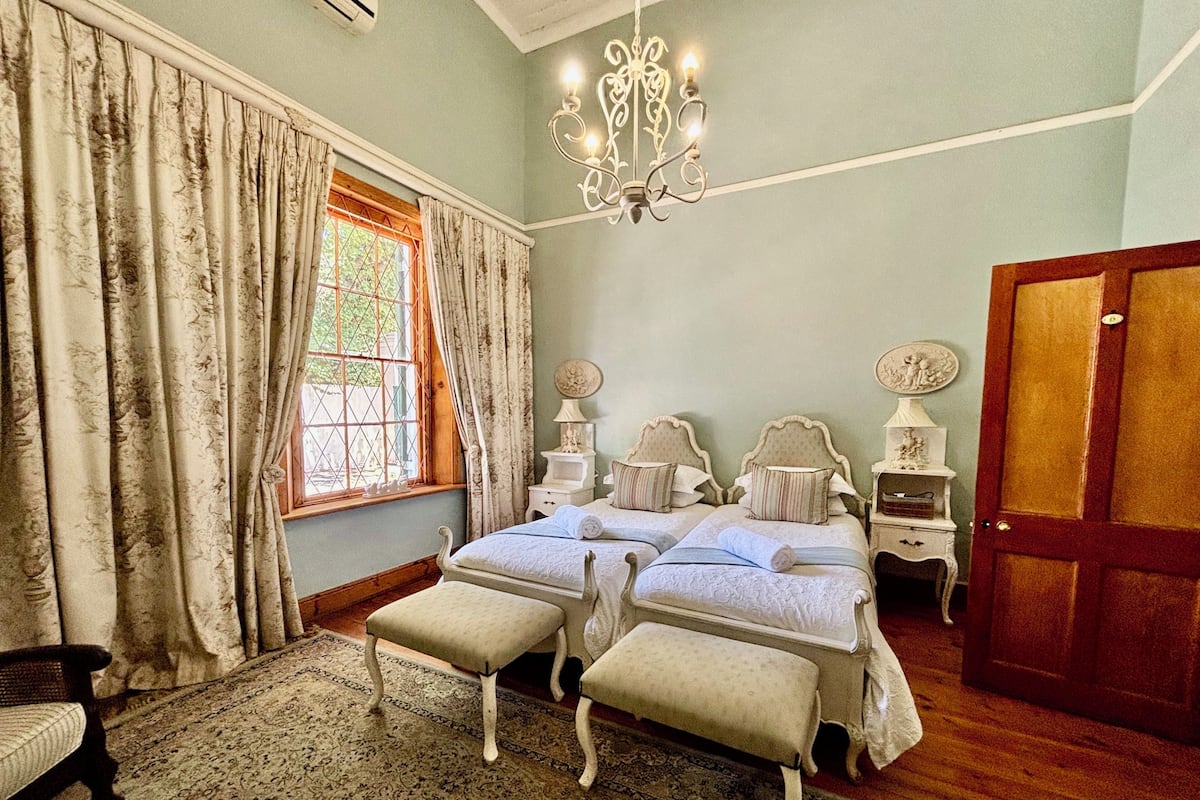 A bedroom with two beds, a chandelier, a wooden floor, and a large window with curtains.