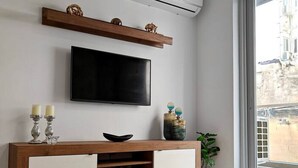 A modern living room with a flat-screen TV mounted on the wall, a wooden console table with decorative items, a potted plant, and a large window providing a view of the outside. - Charming 1-Bedroom Apartment in Il-Gżira, Malta (Il-Gżira)