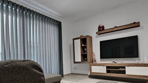 Ein modernes Wohnzimmer mit einem grauen Sofa, einem Flachbildfernseher auf einem Holzboden, einem Spiegel und einem Fenster mit grauen Vorhängen.