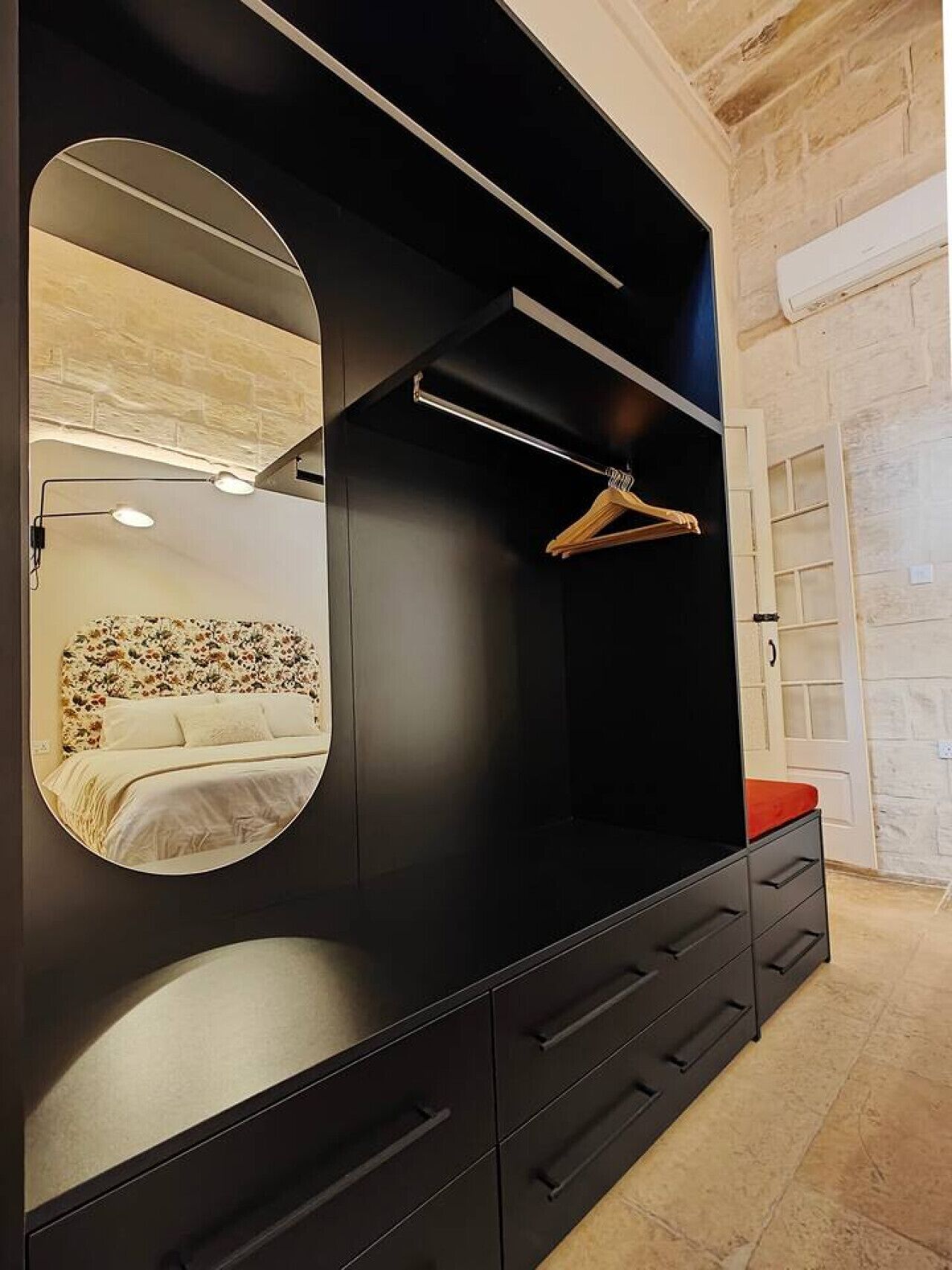 Une chambre moderne avec un grand miroir, un lit doté d’une tête de lit à motifs floraux et une armoire noire avec des tiroirs et un espace pour suspendre les vêtements.