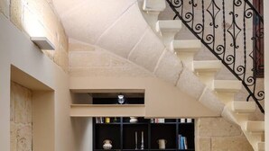 Un couloir étroit avec un escalier menant à un palier. L’escalier est doté d’une rampe en fer forgé noir et le sol est recouvert d’une moquette beige. Sur le palier, on trouve une bibliothèque remplie de livres et d’objets décoratifs.