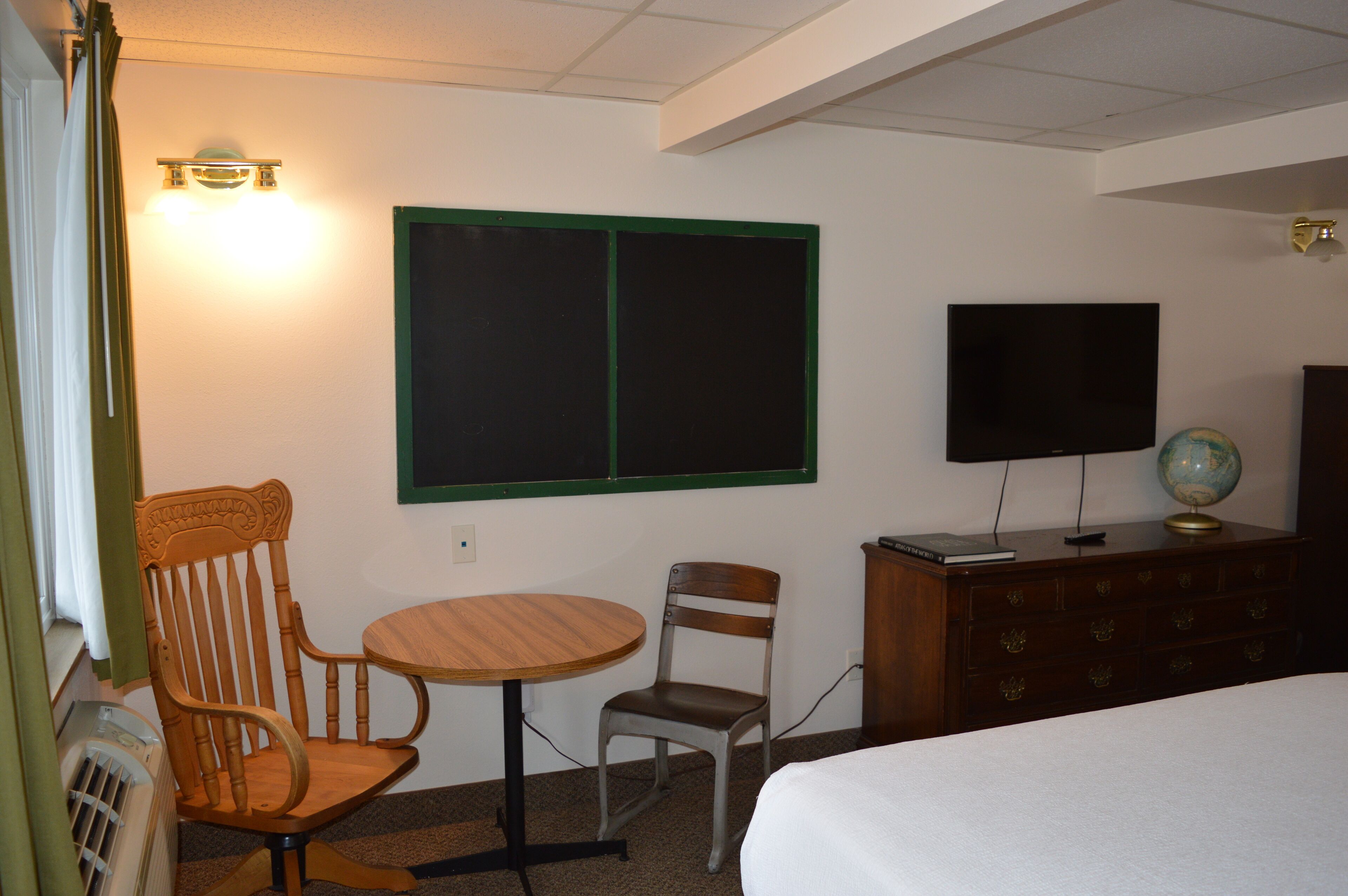 A hotel room with a bed, a wooden chair, a small round table, a television, a globe, and a dresser.