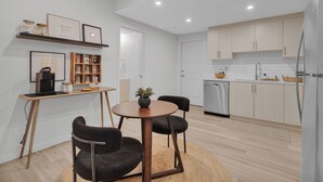 Een moderne keuken met houten kastjes, een inbouwvaatwasser en een koffieapparaat. Er is een eethoek met een ronde tafel en twee stoelen.