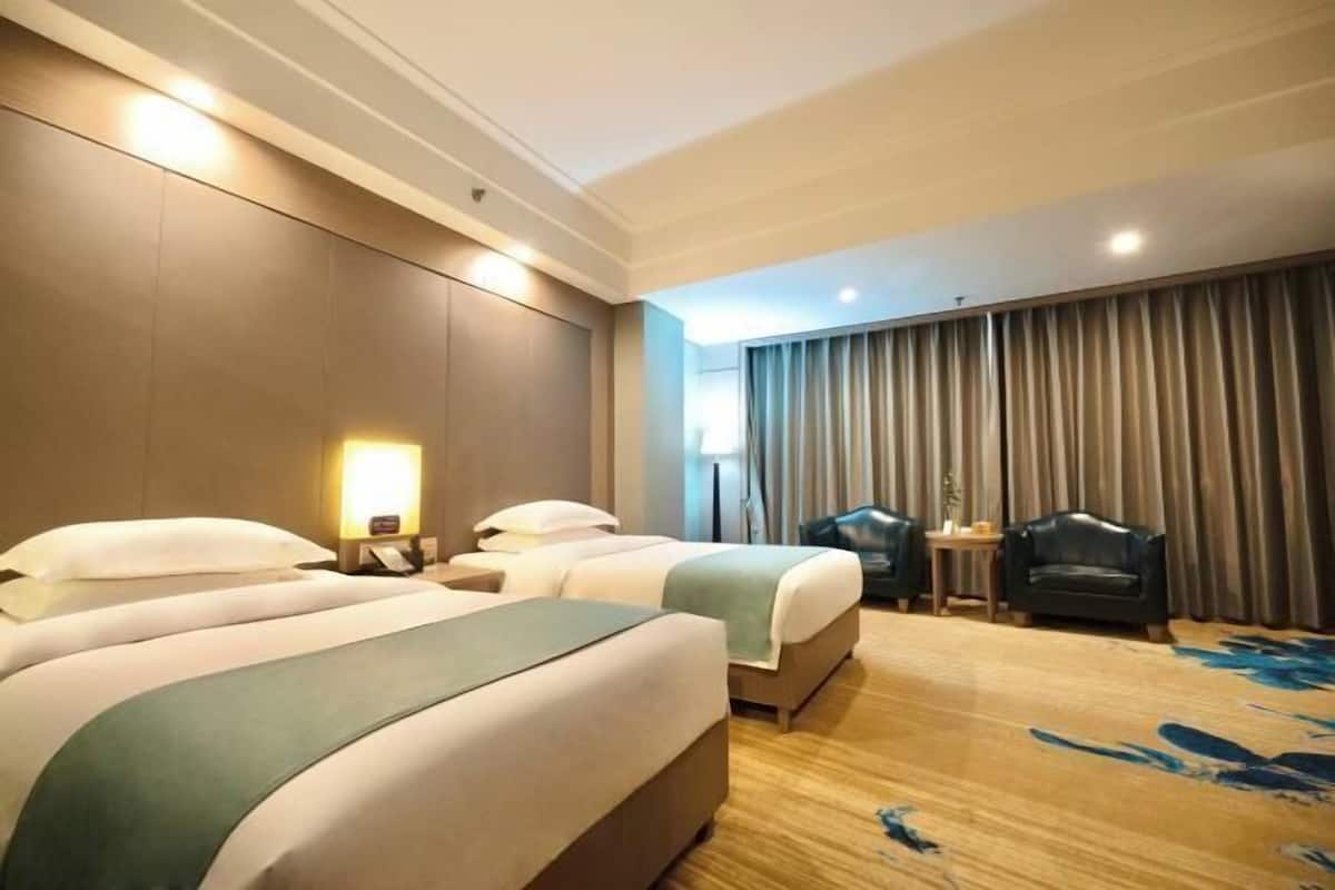 Habitación de hotel con dos camas, un escritorio, una silla y un amplio ventanal con cortinas.