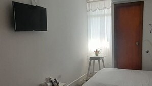 A hotel room with two beds, a television mounted on the wall, a small table with a cup and a bottle, and a window with curtains. - HOTEL HABIBI (Barranquilla)