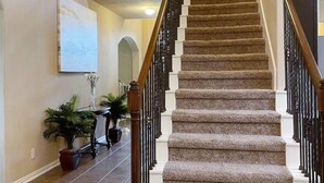 Interior de una casa de varios niveles, con escalera alfombrada, barandillas de madera y plantas en macetas.
