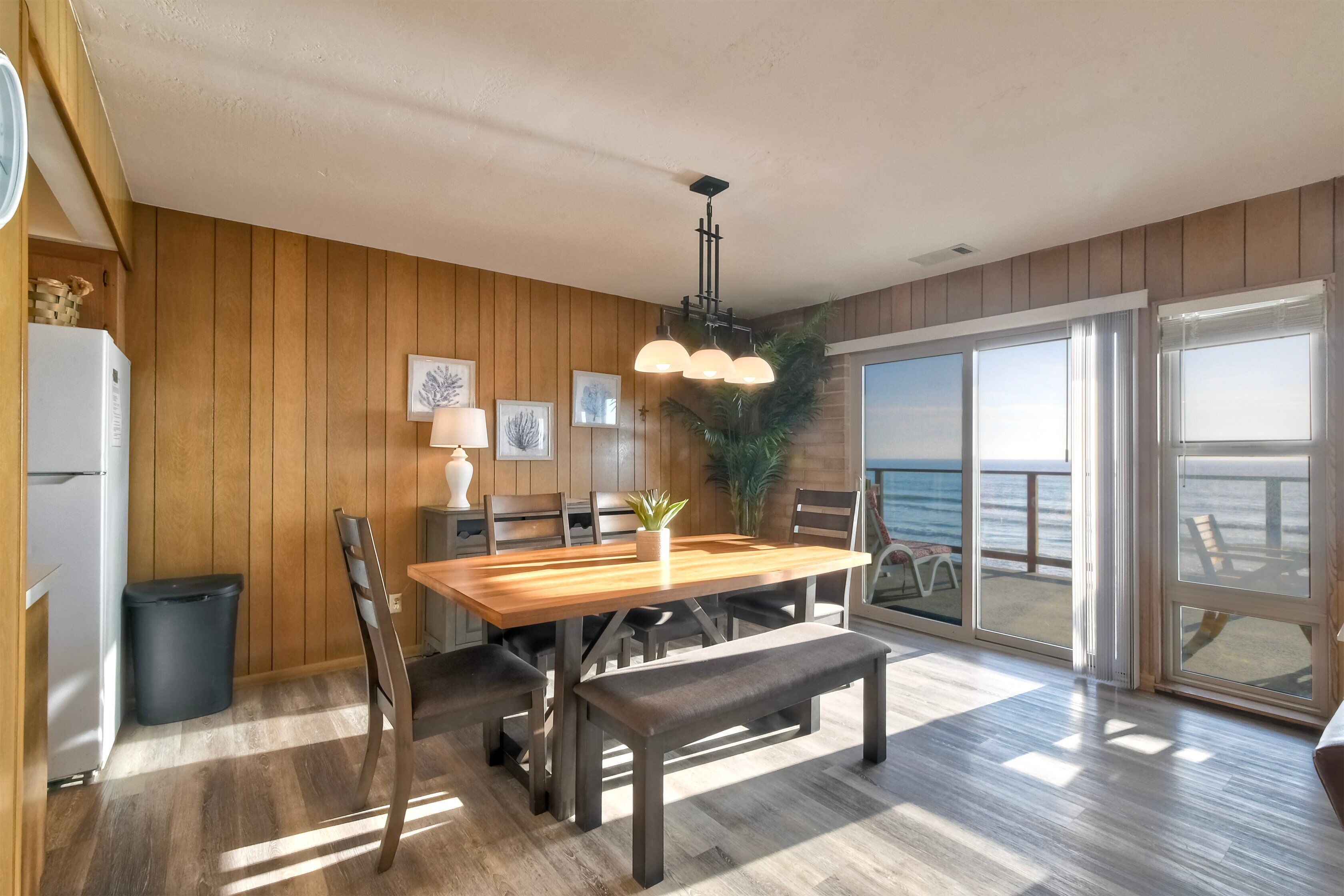 A spacious dining area with a wooden table, benches, and a view of the ocean through large sliding glass doors.