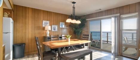 A spacious dining area with a wooden table, benches, and a view of the ocean through large sliding glass doors.