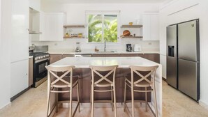 A modern kitchen with white cabinets, a central island with bar stools, a large refrigerator, and a window with a view of palm trees. - Private 3BR Villa with Pool, BBQ & Spacious Garden (Punta Cana)