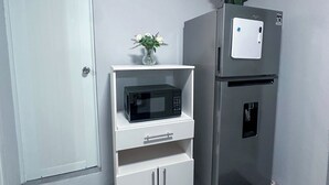 A compact kitchen with a white microwave on a white cabinet, a stainless steel refrigerator, and a small white shelf with a vase of yellow flowers.