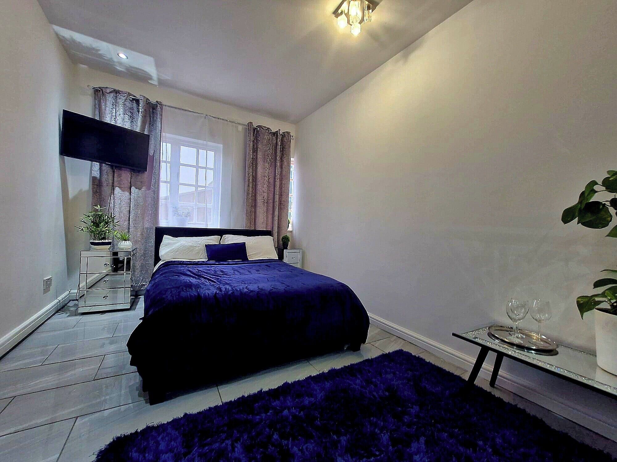 A bedroom with a bed, a bedside table with a lamp, a window with curtains, and a small table with glasses.