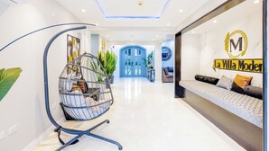 Un hall d’hôtel moderne avec un plafond incurvé, un fauteuil suspendu agrémenté de plantes, un canapé orné d’un logo et, au fond, une porte bleue.