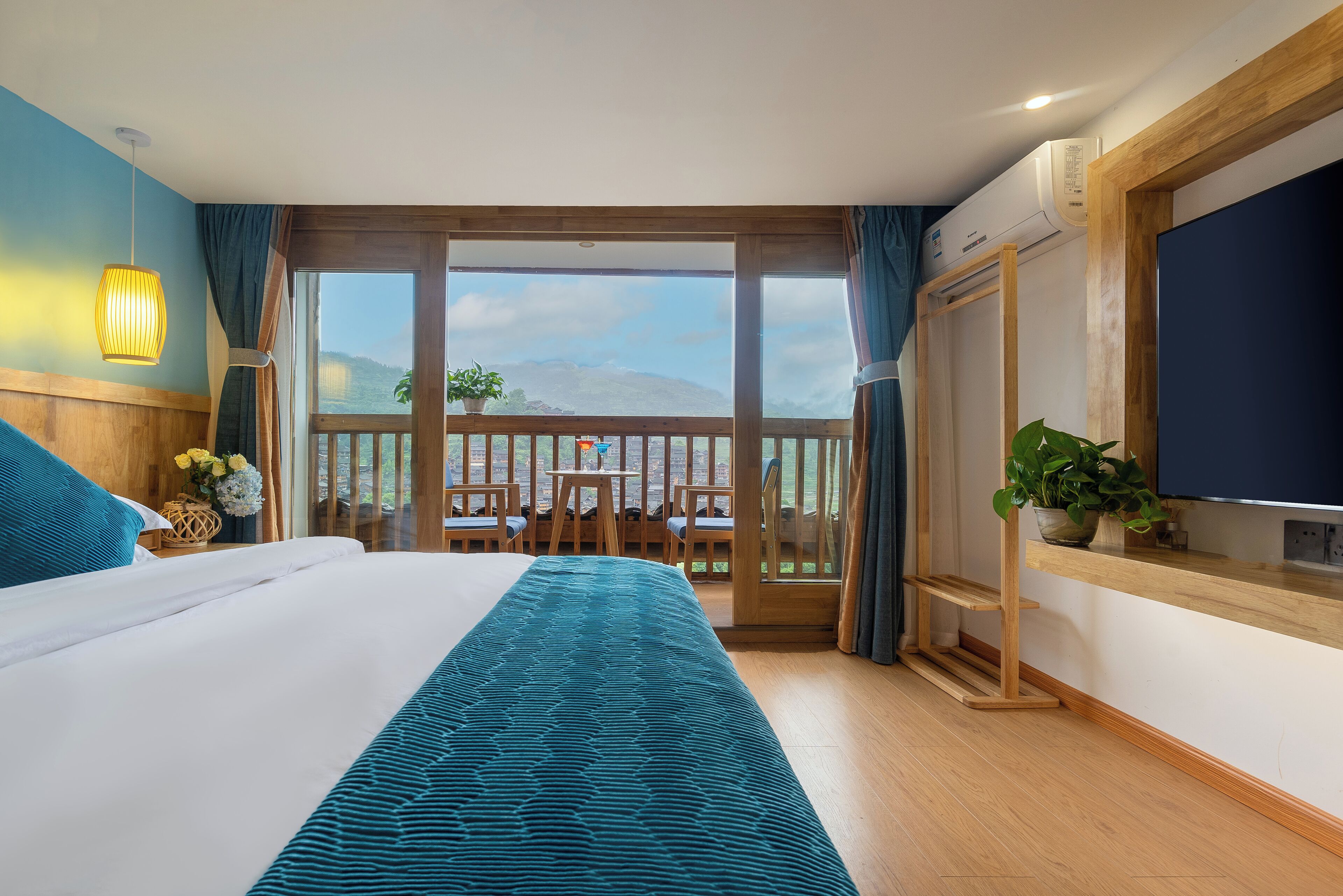 A hotel room with a large bed, a balcony with a wooden railing, a TV, and a potted plant.