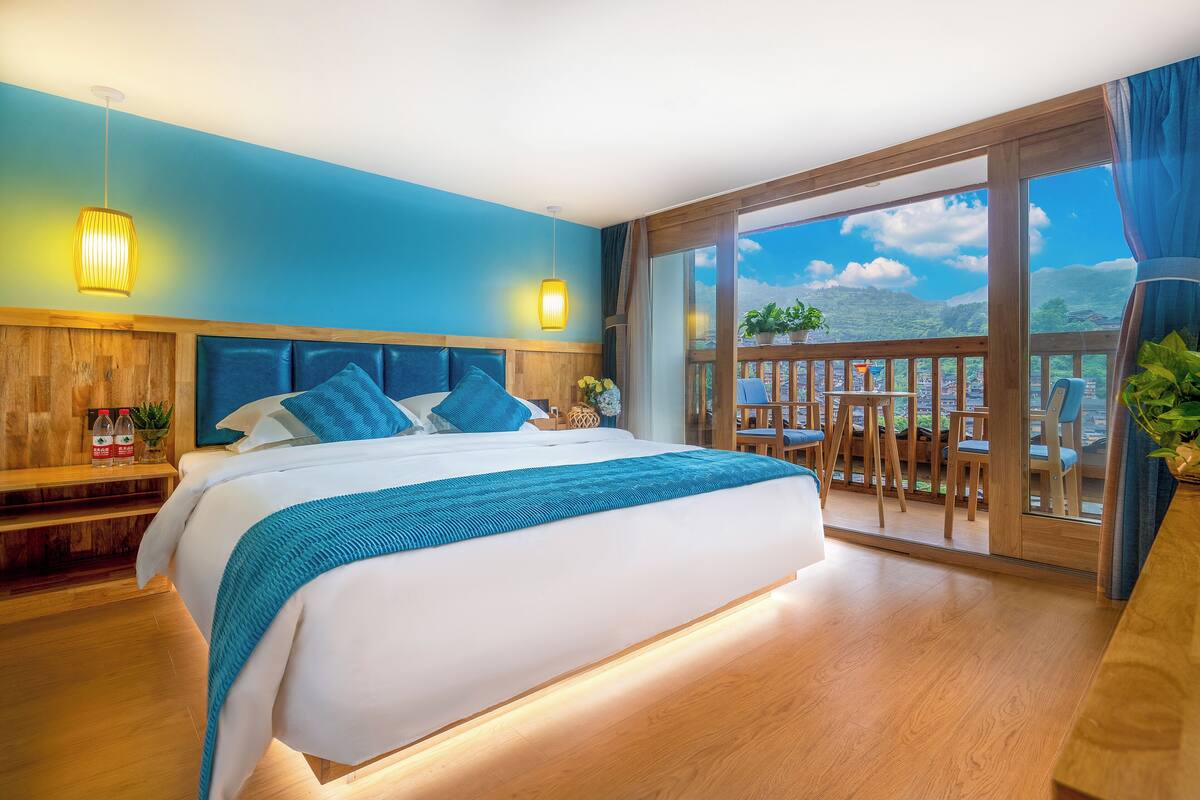 A bedroom with a large bed, wooden headboard, and a balcony with a view of the city and mountains.