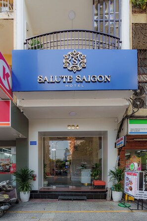 The entrance of the Salute Saigon Hotel with a blue sign, a glass door, and potted plants in front.
