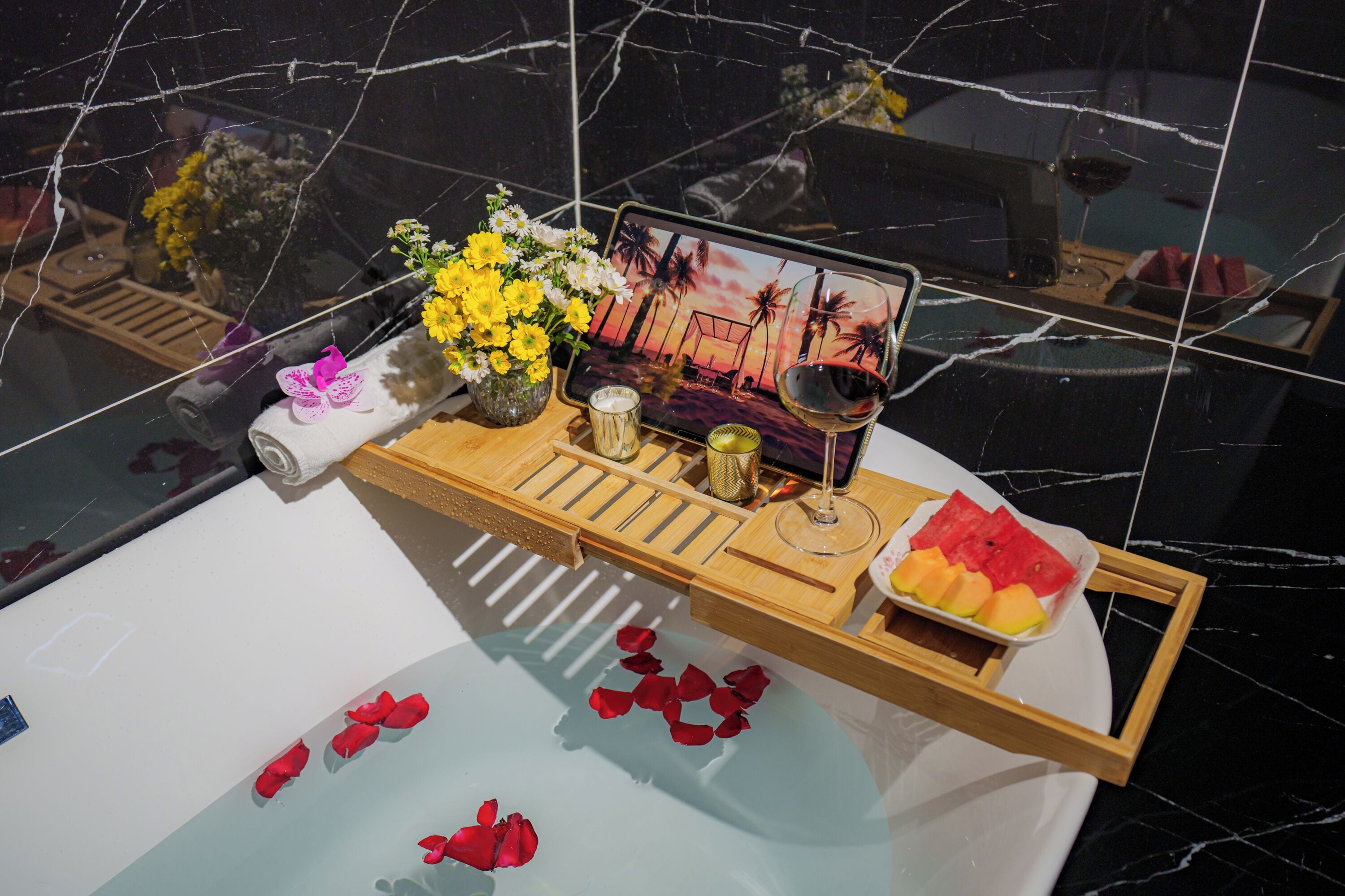 Un tocador de baño con espejo, una bandeja con una copa de vino, flores y fruta.