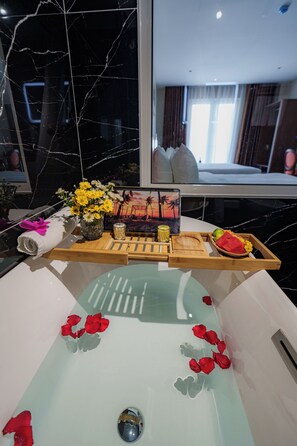 A bathtub with rose petals, a tray with fruits and flowers, and a painting of a sunset.