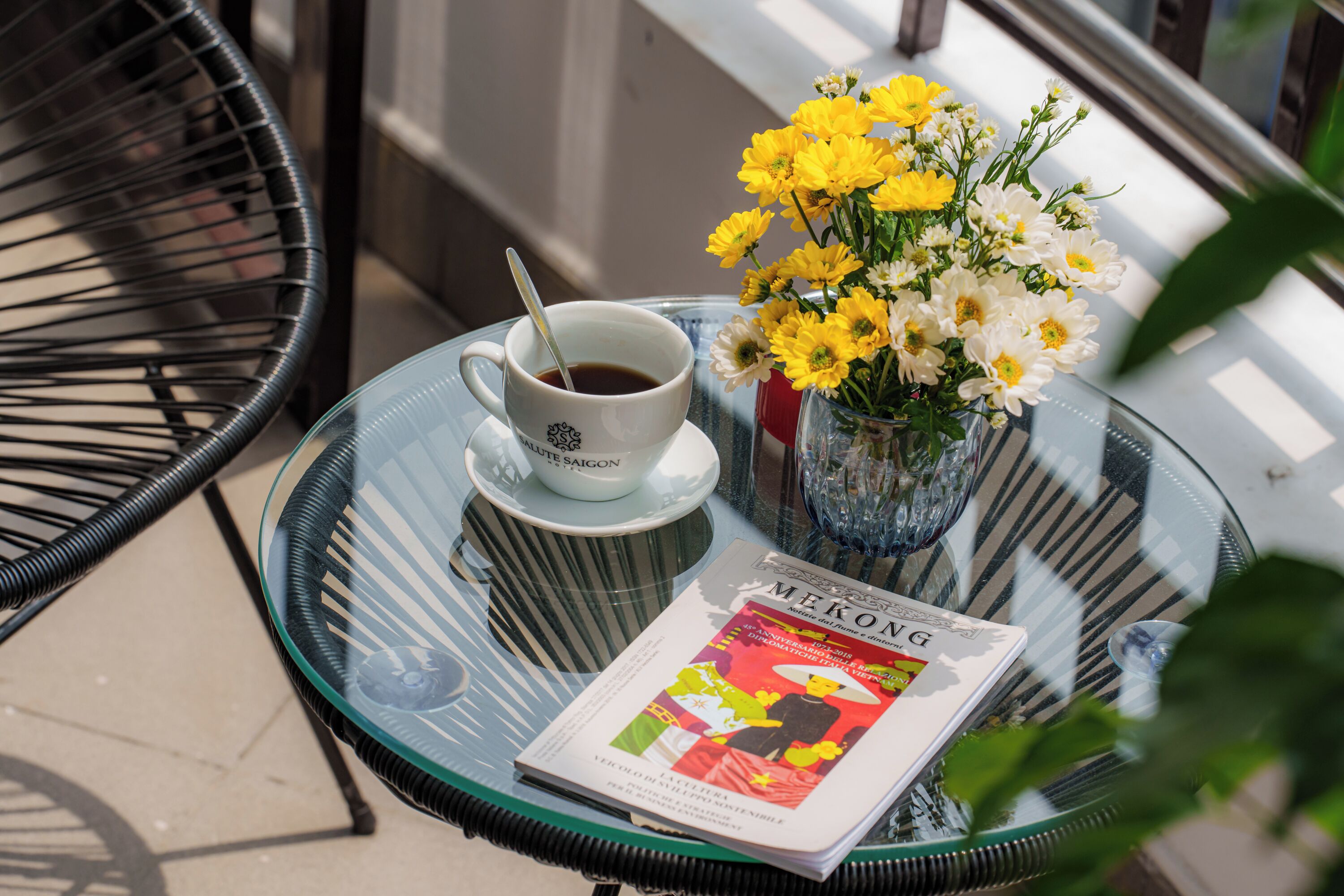 A glass table with a cup of coffee, a vase of yellow flowers, and a magazine.