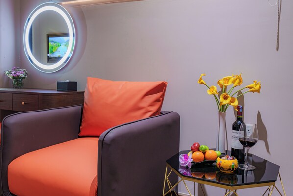 A modern room with a comfortable orange chair, a small table with a vase of yellow flowers, a bottle of wine, and a fruit bowl.