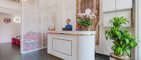 A reception desk with a receptionist, a decorative wall, a potted plant, and a seating area with red chairs.