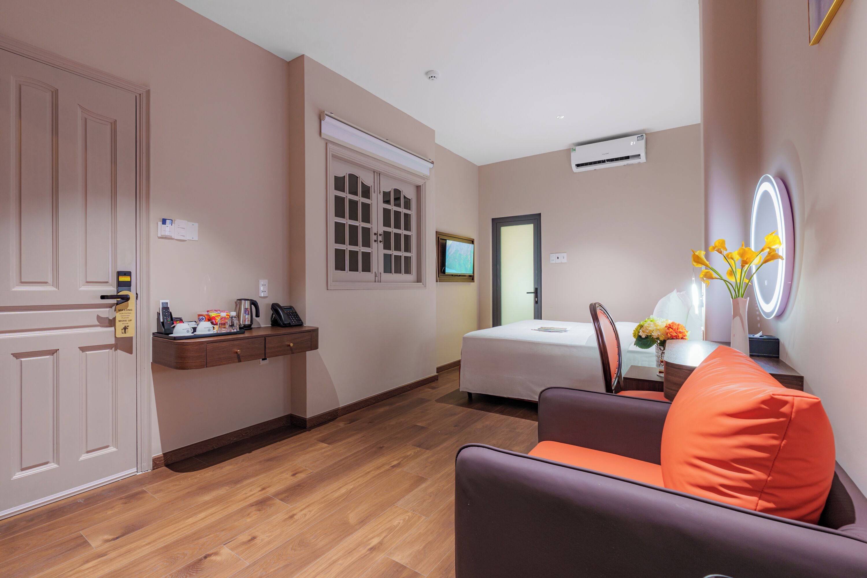 Habitación de hotel moderna con piso de madera, un sofá con un cojín naranja, una mesita de noche con un florero, una cama, un televisor y una puerta con un cartel.