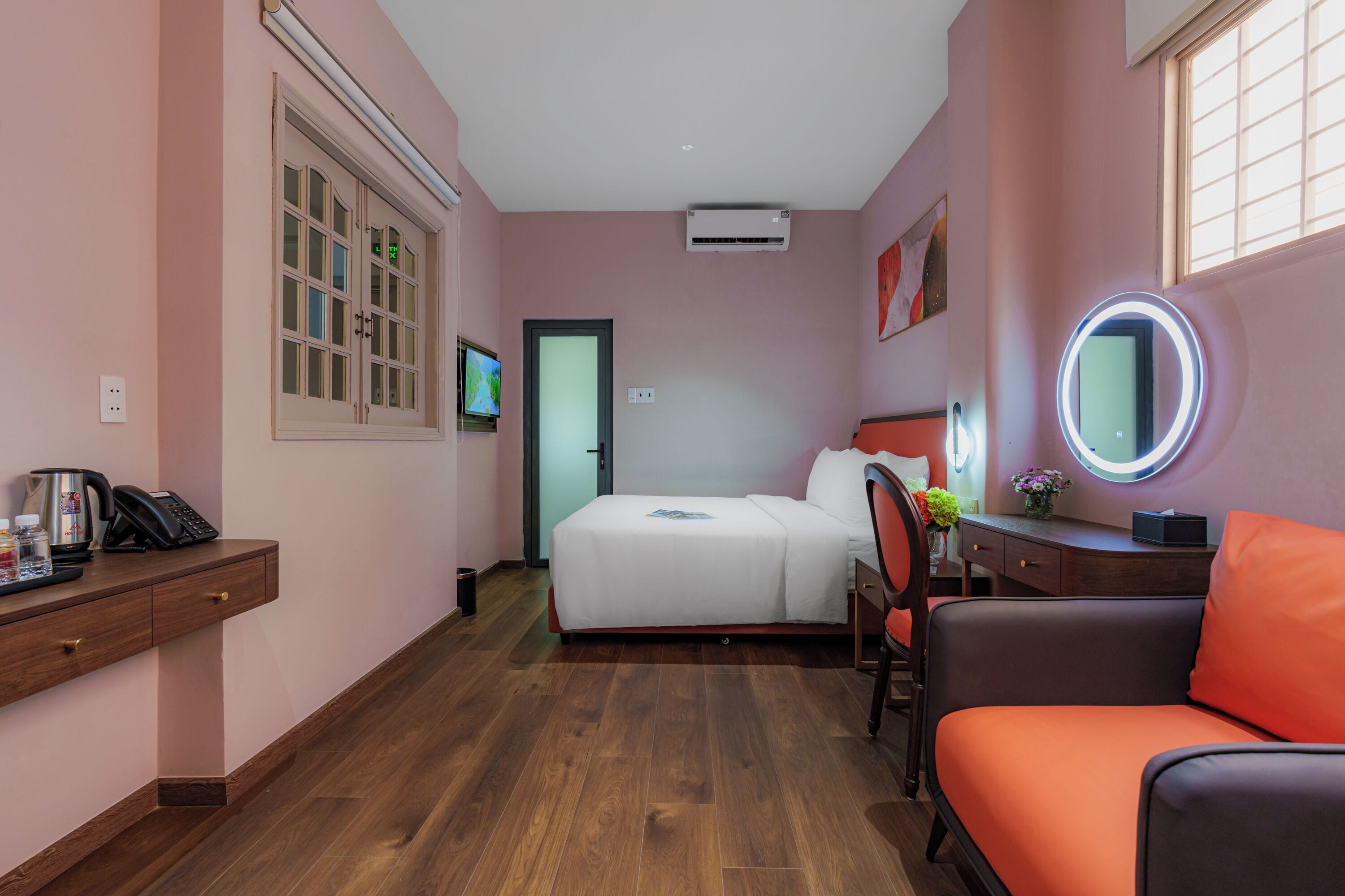 A hotel room with a bed, a desk with a telephone, a chair, a round mirror, and a window with blinds.