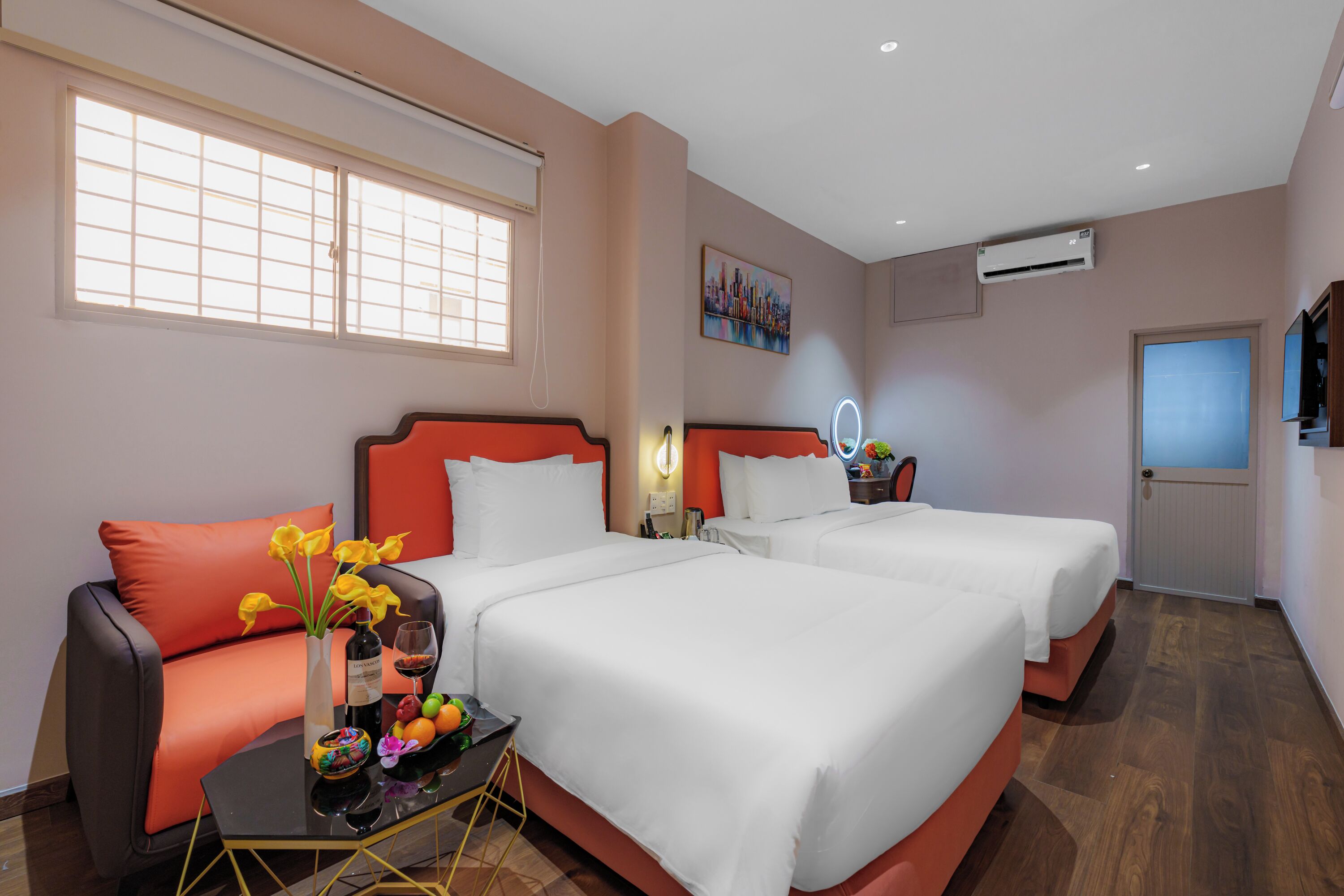 Habitación de hotel con dos camas, un sofá, una mesita con un florero y una ventana con persianas.