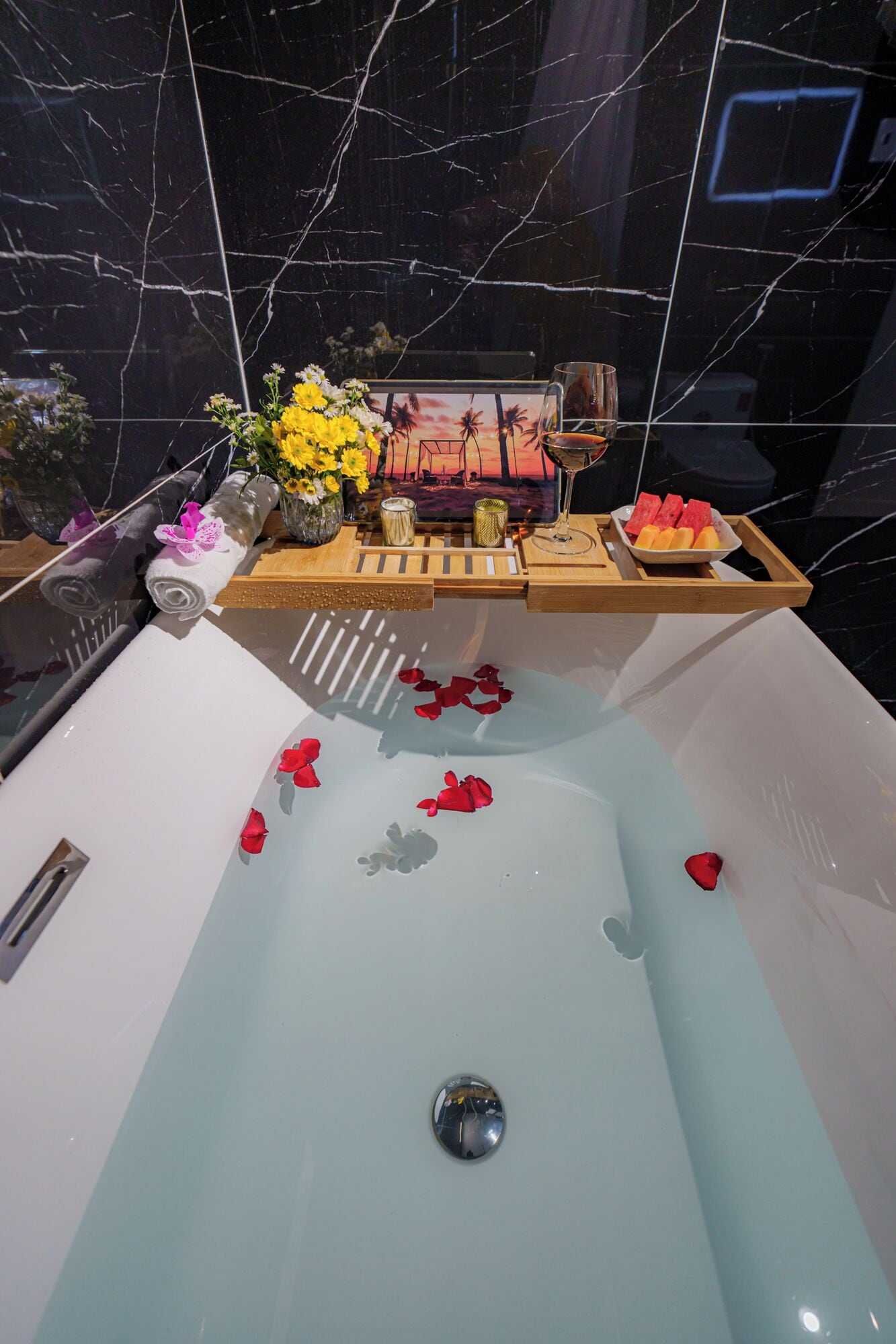 A bathtub with rose petals, a tray with fruits, and a decorative item on a wooden surface.