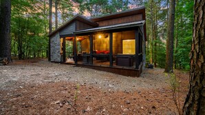 Une cabane aux murs en pierre, dotée d’un porche abrité et d’une grande fenêtre donnant sur une forêt.