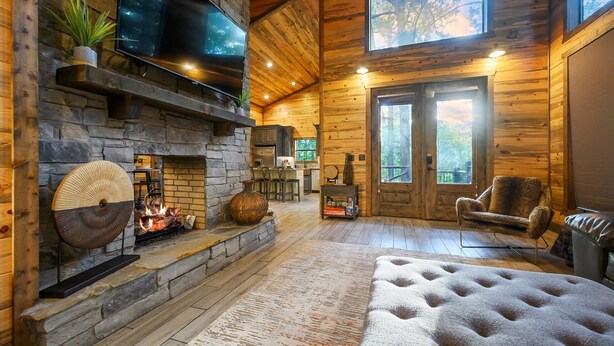 L’intérieur chaleureux d’un chalet avec une cheminée en pierre, un téléviseur fixé au mur et une grande fenêtre laissant entrer la lumière naturelle.
