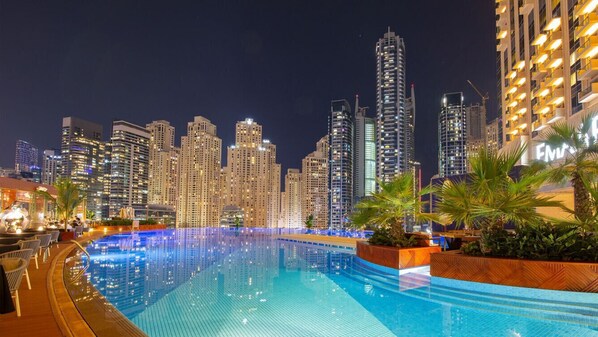 A nighttime cityscape with a pool area featuring a city view, palm trees, and a well-lit building.